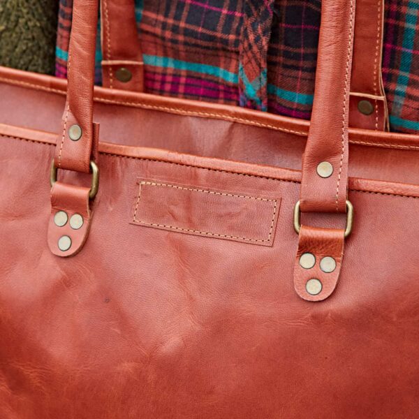 Close-up of a Personalised Brown Leather Zipped Tote Shopping Bag (Copy) with brass hardware, worn over the shoulder of someone in a checked shirt with red, green, and blue tones.