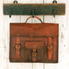 The Personalised Brown Leather Satchel (Copy) with buckles and a front pocket hangs from a carved wooden wall hook against a white wooden background.