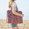 A person in a pink T-shirt and denim shorts stands on a pebbled beach carrying a watermelon quilted cotton tote bag.