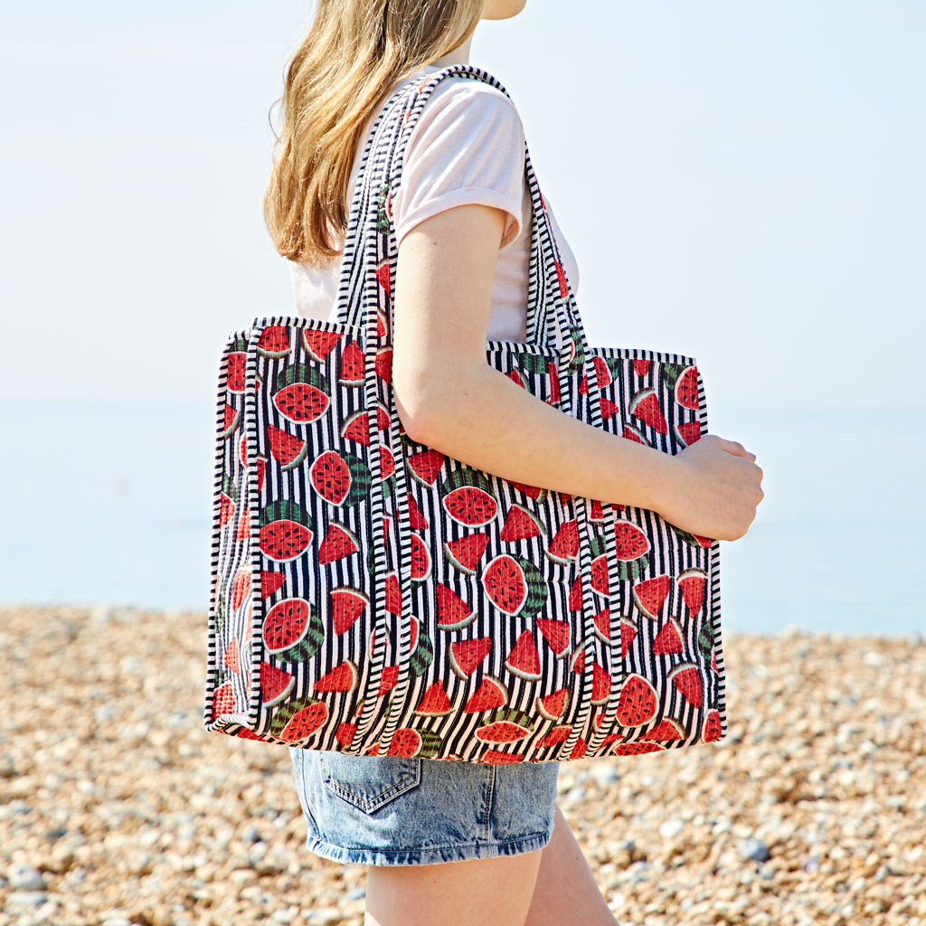 A person in a pink T-shirt and denim shorts stands on a pebbled beach carrying a watermelon quilted cotton tote bag.