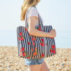 A person in a white T-shirt and denim shorts holds a strawberry quilted cotton tote bag, standing on a pebbled beach near the sea.
