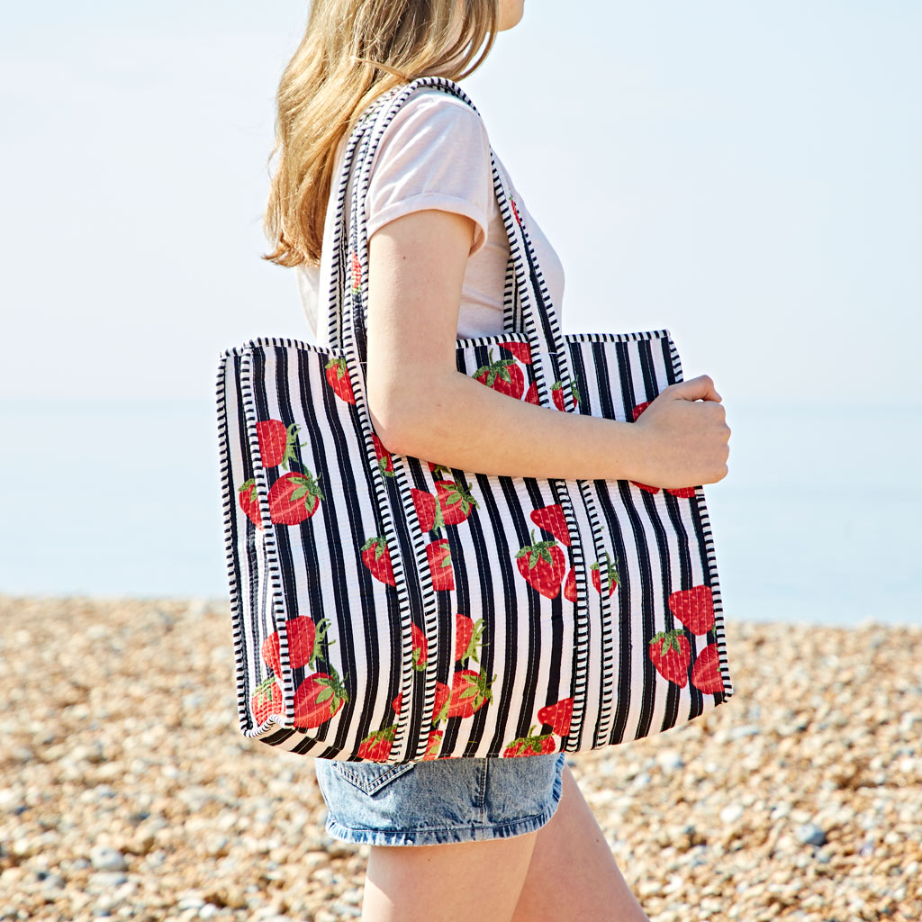 A person in a white T-shirt and denim shorts holds a strawberry quilted cotton tote bag, standing on a pebbled beach near the sea.