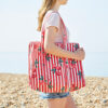 A person wearing a light pink shirt and denim shorts carries a red-and-white striped tote bag with a cherry pattern whilst standing on a pebbled beach.
