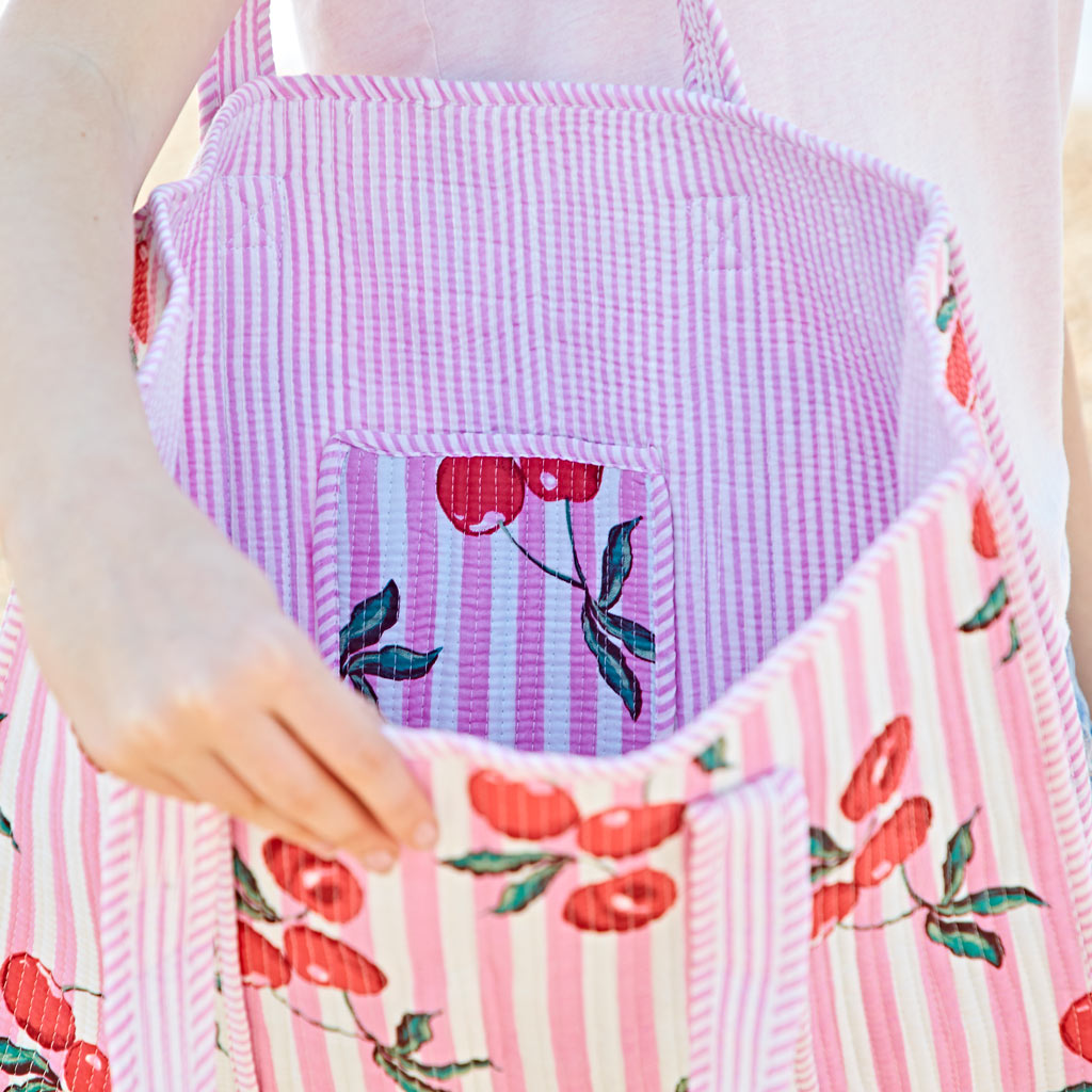 A person holds open a pink and white striped tote bag with a cherry pattern, showing the interior and a matching inside pocket.