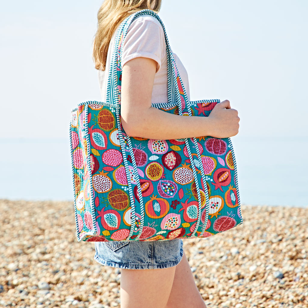 A person standing on a pebble beach carries a colourful, patterned tote bag over their shoulder, wearing a white T-shirt and denim shorts.