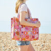 A person in a light pink T-shirt and denim shorts carries a pomegranate quilted cotton tote bag on a pebbled beach.
