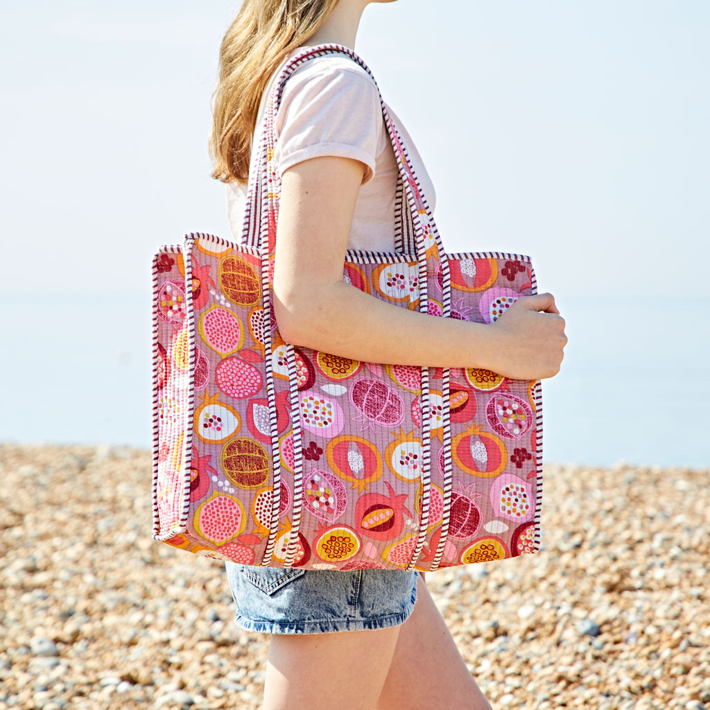 A person in a light pink T-shirt and denim shorts carries a pomegranate quilted cotton tote bag on a pebbled beach.
