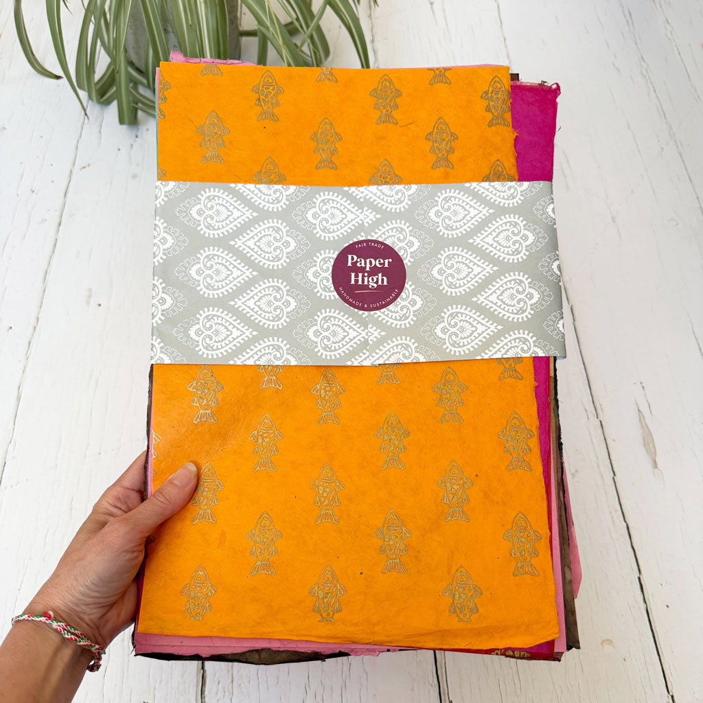 A hand holds the Handmade Lokta Paper Craft Pack against a white wooden background, with a plant visible at the top. The pack displays vibrant patterned paper, ideal for creative projects.