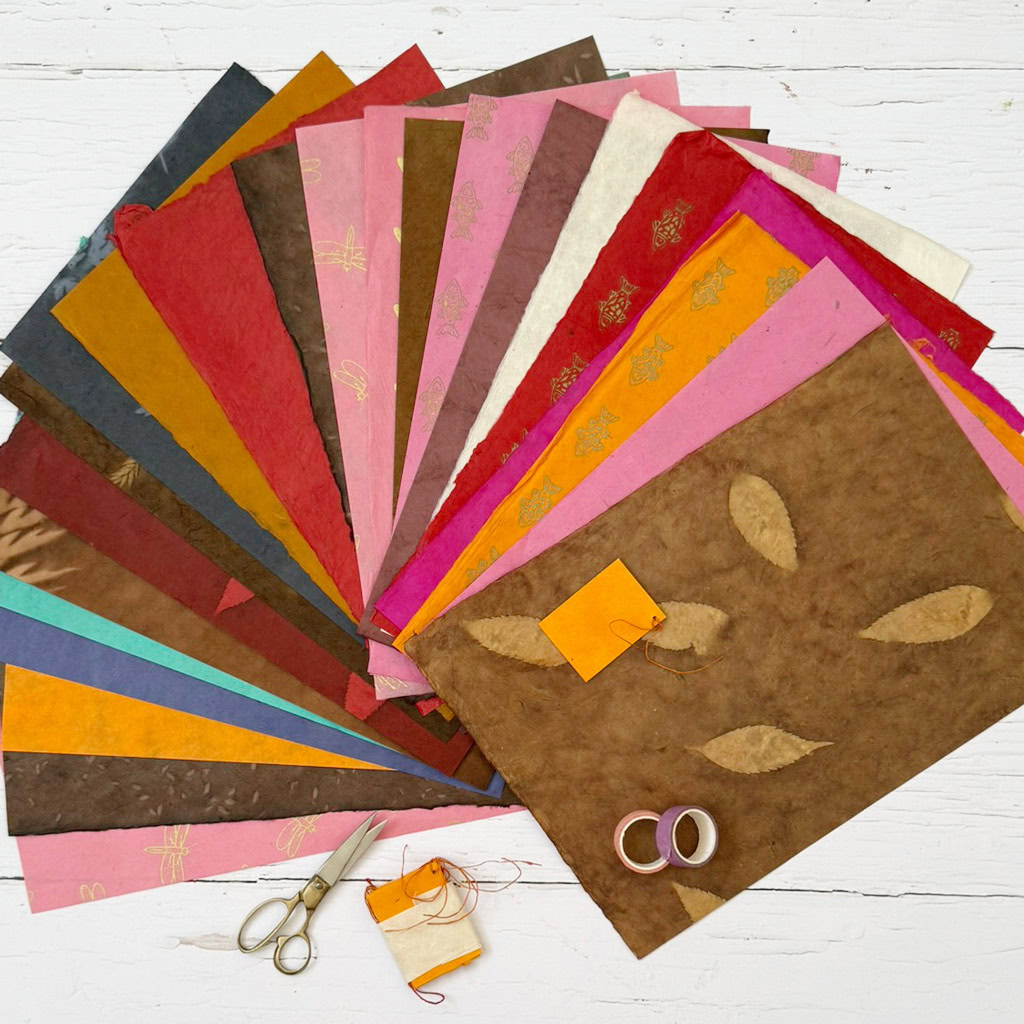 Sheets of colourful decorative paper from the Handmade Lokta Paper Craft Pack are fanned out on a white background, accompanied by scissors, twine, and rolls of washi tape placed alongside.