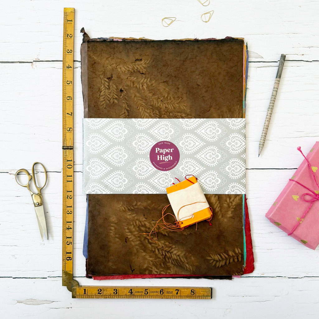 A Handmade Lokta Paper Craft Pack is displayed on a white wooden surface, accompanied by a ruler, scissors, measuring tape, paper clips, and a wrapped present.