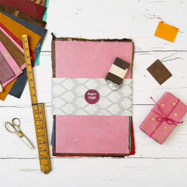 A stack of vibrant sheets from the Handmade Lokta Paper Craft Pack, accompanied by a pink-wrapped present, ruler, scissors, and small brown labels, are neatly arranged on a white wooden surface.