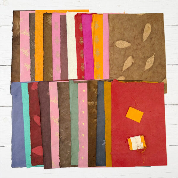 Handmade Lokta Paper Craft Pack featuring assorted colours and textures is arranged in rows on a white wooden surface. Several sheets display patterns such as leaves, hearts, and insects, and two small tags are included.