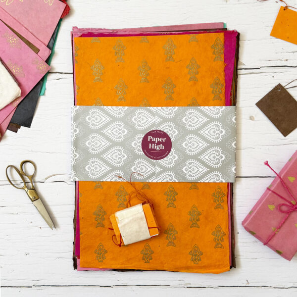 A stack of vibrant, patterned handmade papers from the Handmade Lokta Paper Craft Pack, wrapped with a decorative Paper High label, is displayed on a white wooden surface alongside scissors and small bundles of paper.