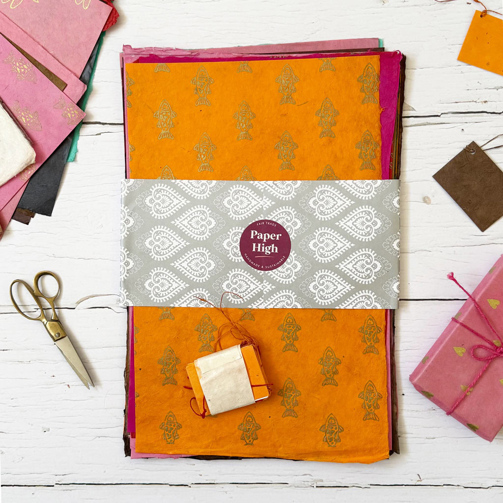 A stack of vibrant, patterned handmade papers from the Handmade Lokta Paper Craft Pack, wrapped with a decorative Paper High label, is displayed on a white wooden surface alongside scissors and small bundles of paper.