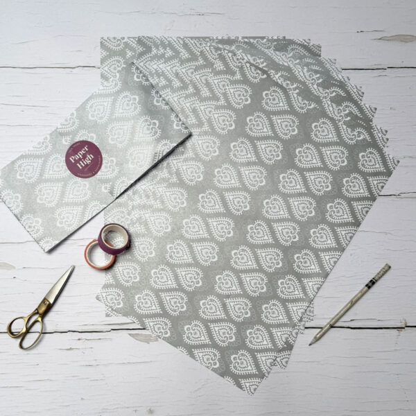 Five sheets of grey patterned paper, a pair of gold scissors, two rolls of tape, a pencil, and an envelope—all included in our Handmade Silver Heart Craft Pack—arranged on a white wood surface.