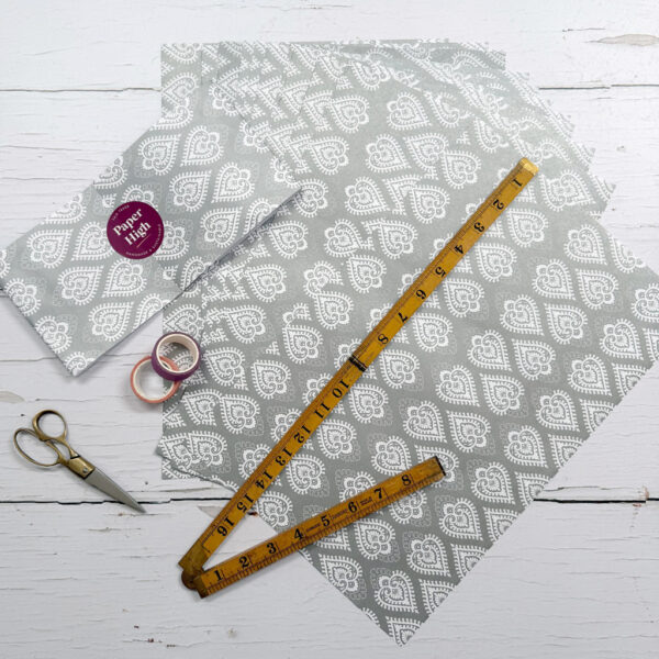 Papers with grey patterns, a folded instruction sheet, a pair of scissors, a folding ruler, and two washi tape rolls from the Handmade Silver Heart Craft Pack are displayed on a white wooden surface.