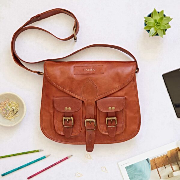 A tan leather satchel personalised with the name "LAURA" sits on a white surface with pencils, a succulent, paper clips, and a mobile phone.